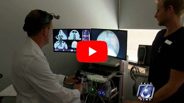 A doctor in a white coat uses medical equipment and monitors showing scan images, while another person stands nearby. A large red play button overlays the center, and a news logo appears in the bottom right corner.