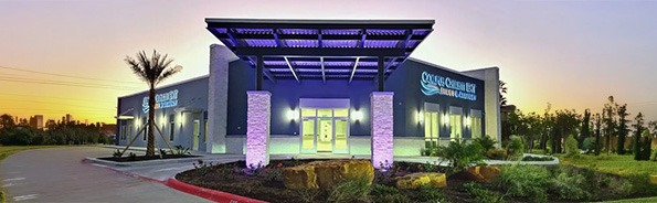 Modern medical building with a covered entrance and purple lighting, surrounded by landscaped plants and rocks, at sunset. The sign on the building reads Sagewell Health & Rehabilitation.