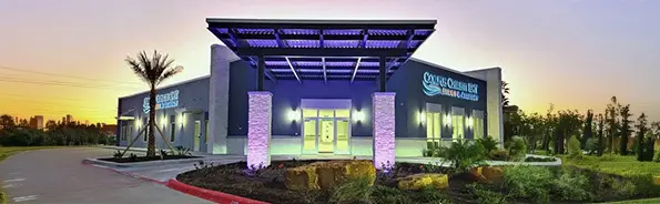 Modern medical building with a covered entrance and purple lighting, surrounded by landscaped plants and rocks, at sunset. The sign on the building reads Sagewell Health & Rehabilitation.