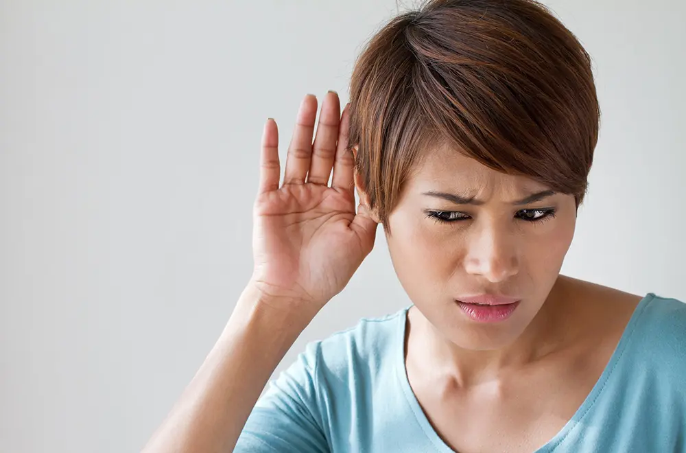 A woman with short brown hair holds her hand to her ear and looks confused or concerned, as if trying to hear something better. She is wearing a light blue top and is set against a plain background.