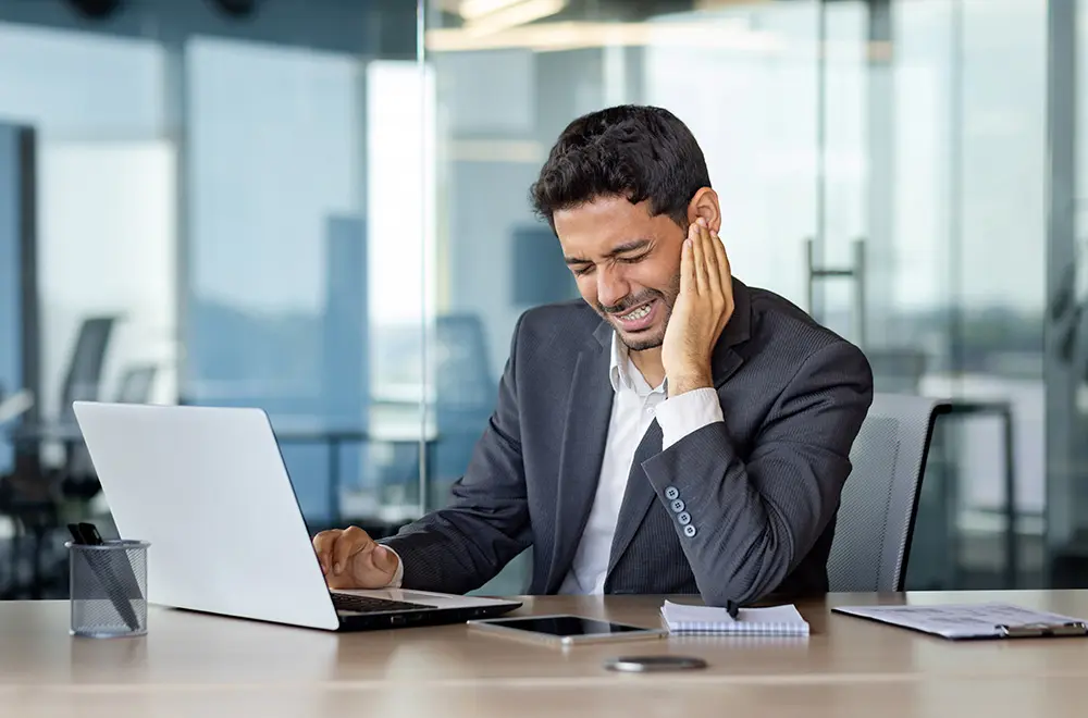 A man in a suit sits at a desk with a laptop, holding his jaw in pain and wincing. He appears to be experiencing discomfort while working in a modern office setting.