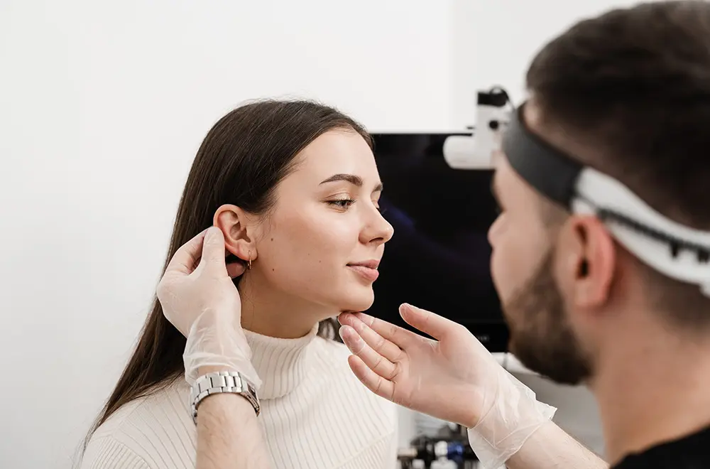 A doctor wearing gloves examines a young woman’s ear as she sits calmly, looking slightly to the side, in a medical office setting.