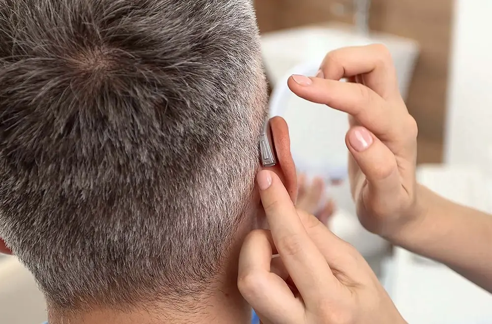 A person fitting a behind-the-ear hearing aid onto the ear of an older adult with short gray hair.