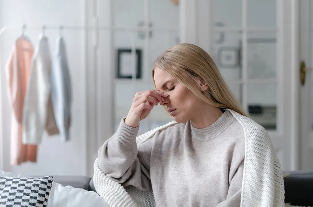 A woman with long blonde hair sits on a couch, eyes closed, pinching the bridge of her nose as if stressed or tired. She wears a light sweater and is draped in a white blanket. Clothes hang in the background.