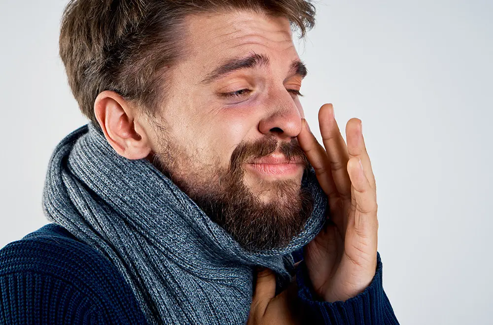 A bearded man wearing a thick gray scarf and blue sweater is closing his eyes and touching his face, appearing to sneeze or feel unwell.