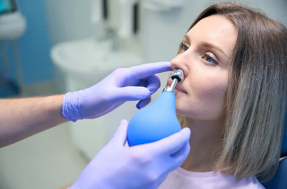 A woman sits while a healthcare professional wearing purple gloves administers nasal inhalation using a blue medical device.