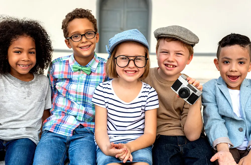 Five smiling children sit close together, dressed in playful, vintage-inspired outfits; one holds a camera, and they appear happy and cheerful outdoors against a blurred white background.