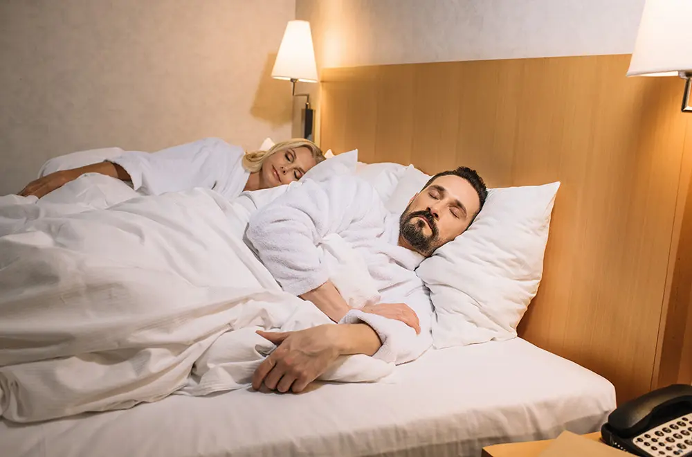 A man and woman wearing white bathrobes are sleeping side by side in a bed with white linens in a softly lit hotel room, with wall-mounted lamps above the headboard.
