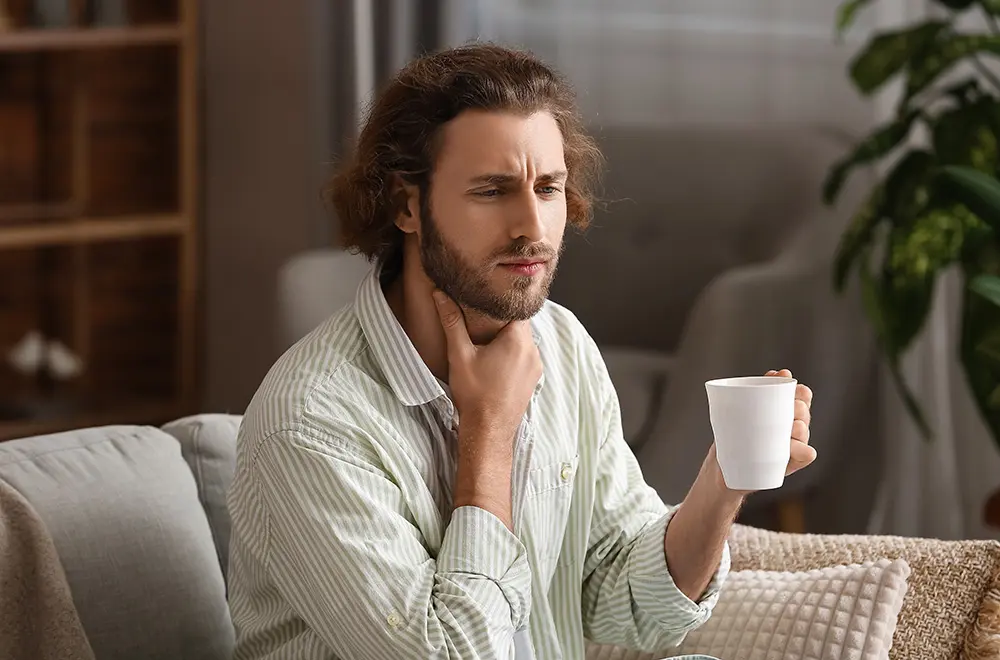 A man sits on a couch holding a white mug in one hand while touching his throat with the other, appearing to be in discomfort or pain, possibly experiencing a sore throat.