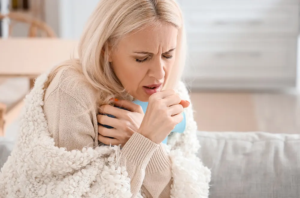 A woman with light hair sits on a couch, wrapped in a blanket, holding a mug and coughing into her fist, appearing unwell.