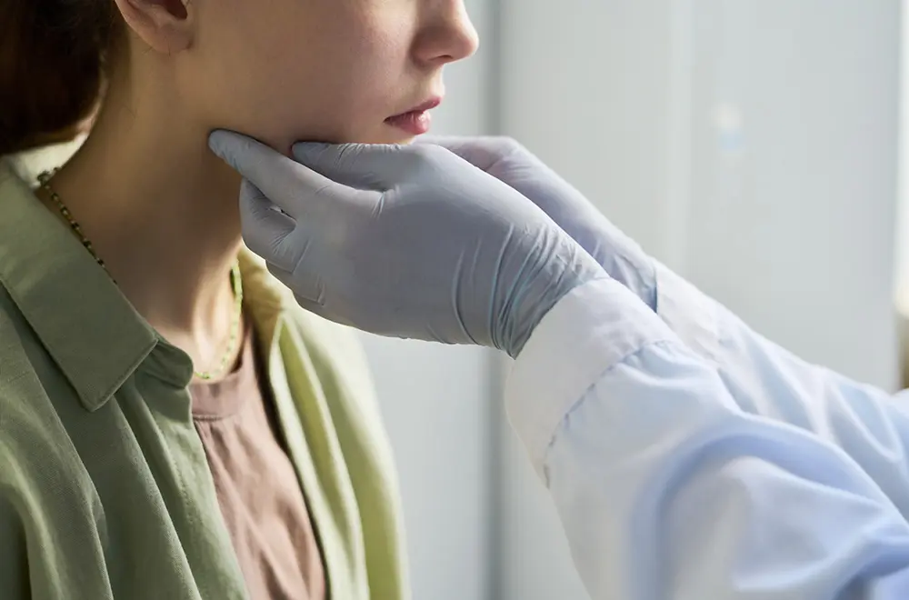 A healthcare professional wearing gloves gently examines a patients neck and jaw. The patient, dressed in a green shirt, sits calmly as the examination takes place in a clinical setting.