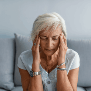 Older woman with short gray hair sits on a couch, eyes closed, pressing her temples with both hands, appearing to have a headache or be stressed. She is wearing a light blue top and silver jewelry.