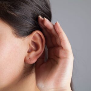A person holds a hand behind their ear, leaning in as if trying to hear better, with close-up focus on the ear and hand against a neutral background.