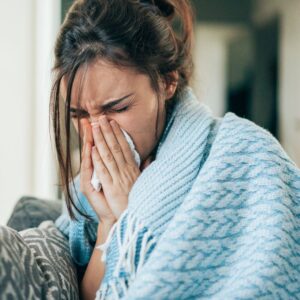 A woman wrapped in a blue blanket sits on a couch, holding a tissue to her nose and appears to be sneezing or coughing.
