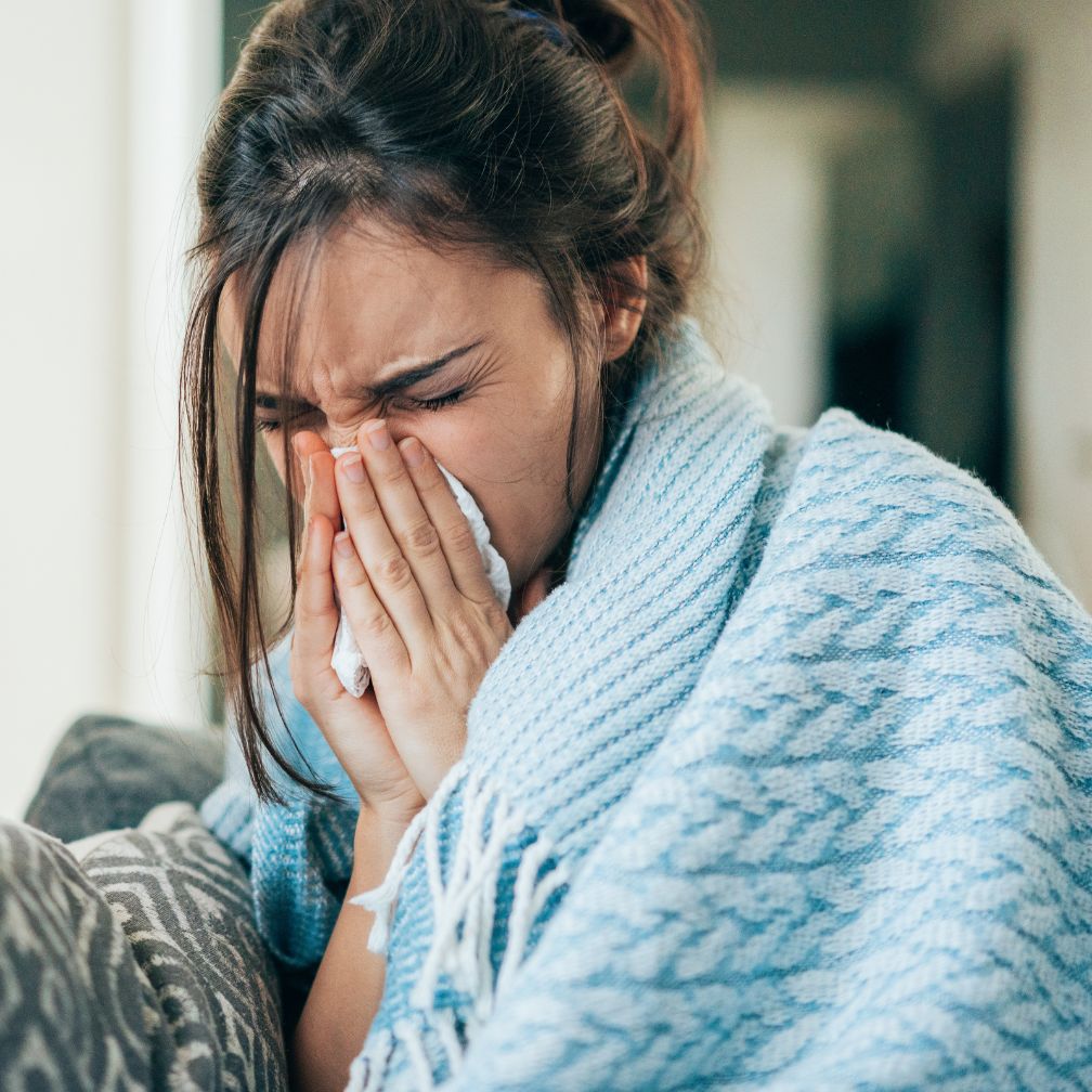 A woman wrapped in a blue blanket sits on a couch, holding a tissue to her nose and appears to be sneezing or coughing.