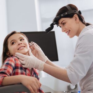 A doctor in a white coat and headlamp examines a young girls ear with an otoscope. The girl, wearing a plaid shirt, sits and smiles while the doctor, wearing gloves, gently checks her ear in a medical office.