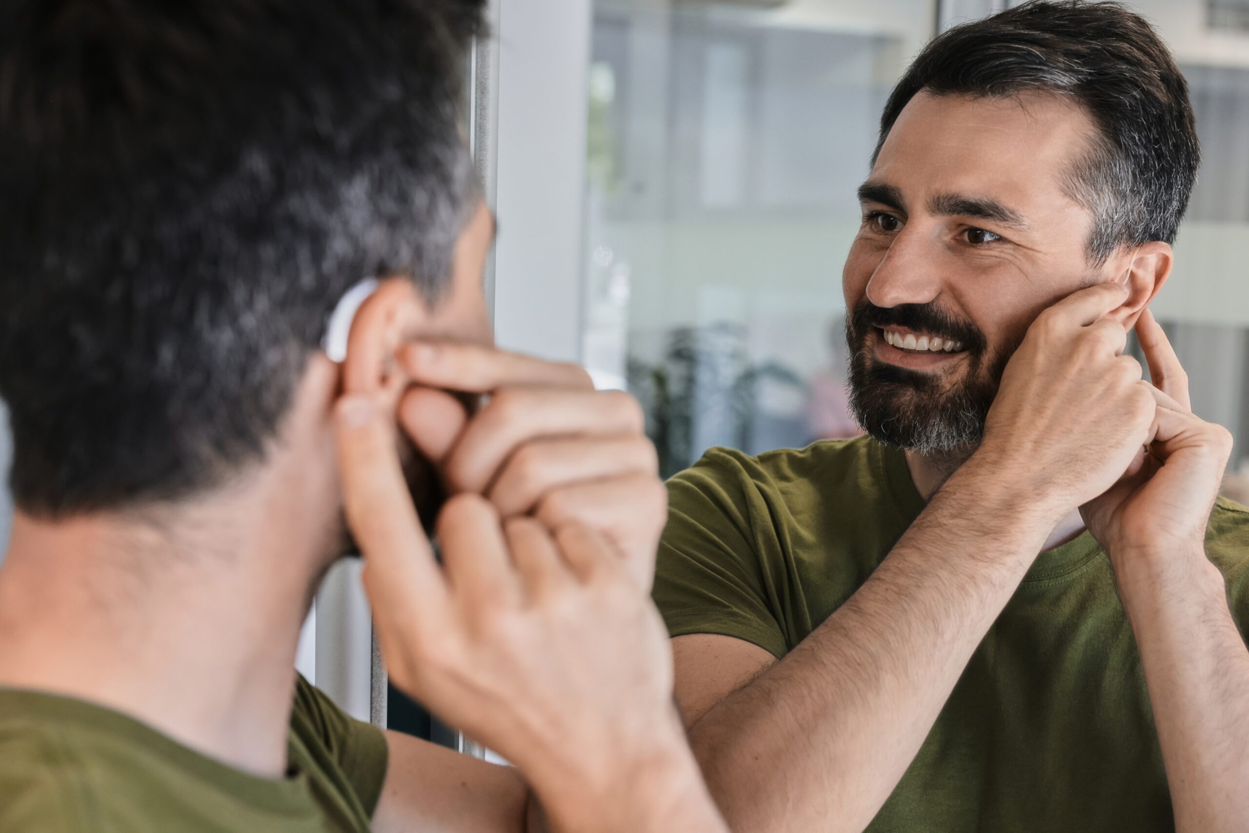 A man with dark hair and a beard smiles while adjusting a hearing aid in his ear, looking at his reflection in a mirror.