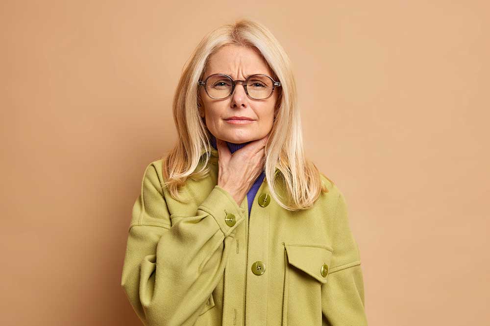 Older woman in glasses and a green jacket touching her throat over a lump-in-the-throat feeling, at Corpus Christi ENT Sinus & Allergy, TX